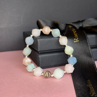 Multicolored beaded bracelet with a gold charm on a pink surface, with 'Ravie Studio' branding.